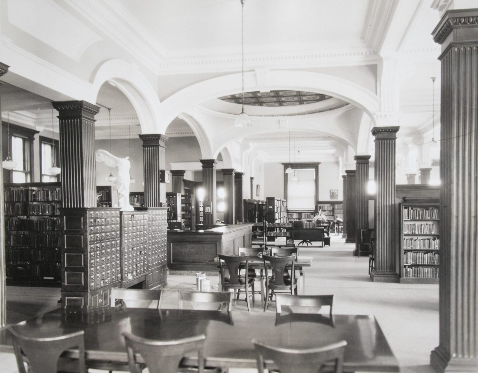 Book shelves, tables and chairs, and card catalogs stand under arched columns in this black and white photo.