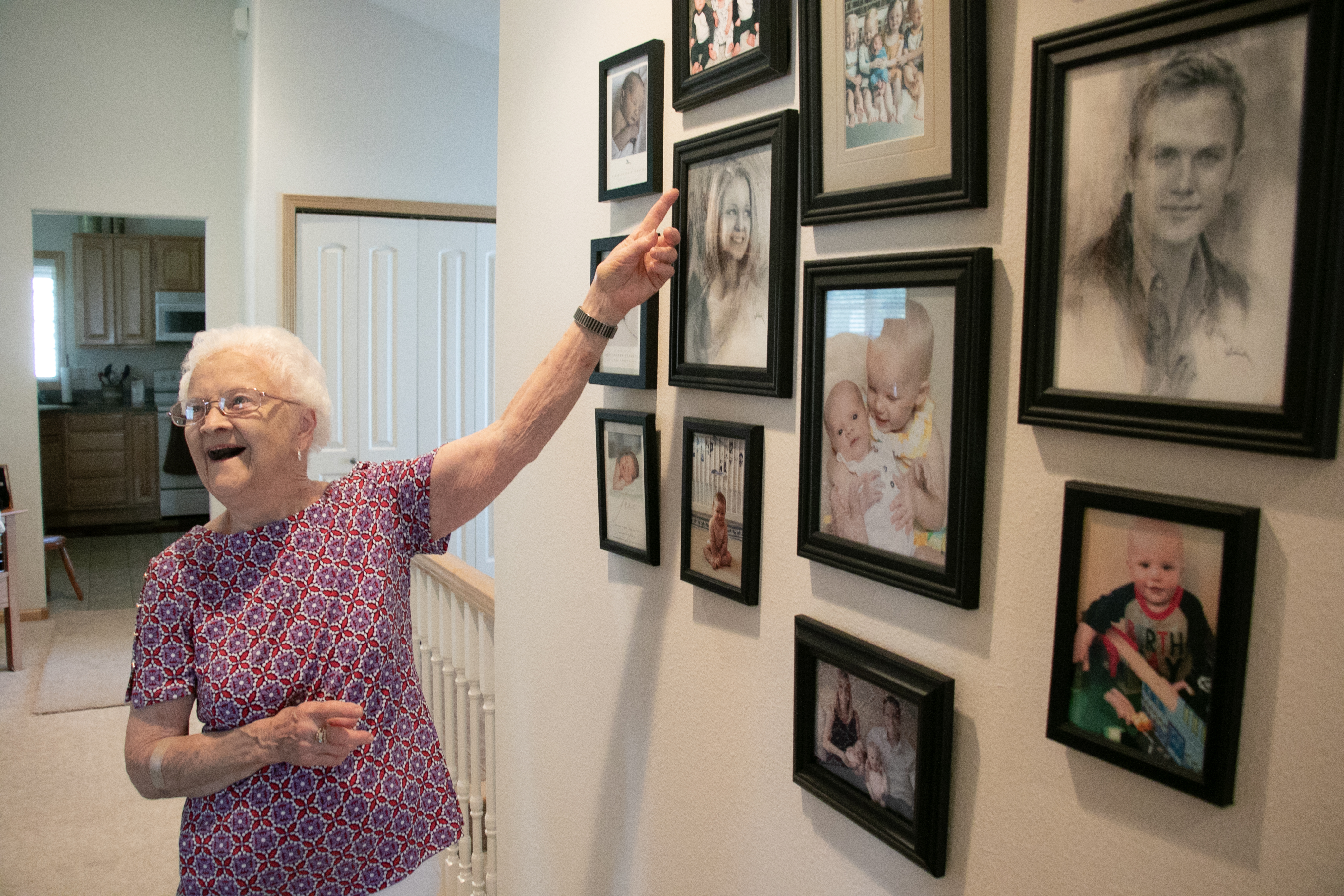 An older woman points to framed photos of her family on her wall and smiles.