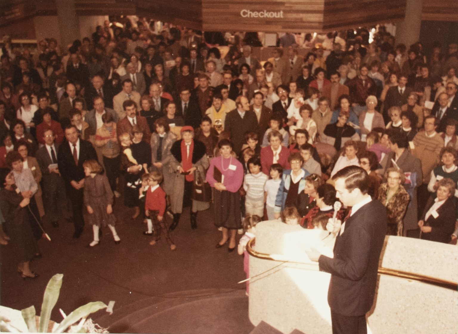 A man in a suit stands on a staircase and speaks while a crowd watches.