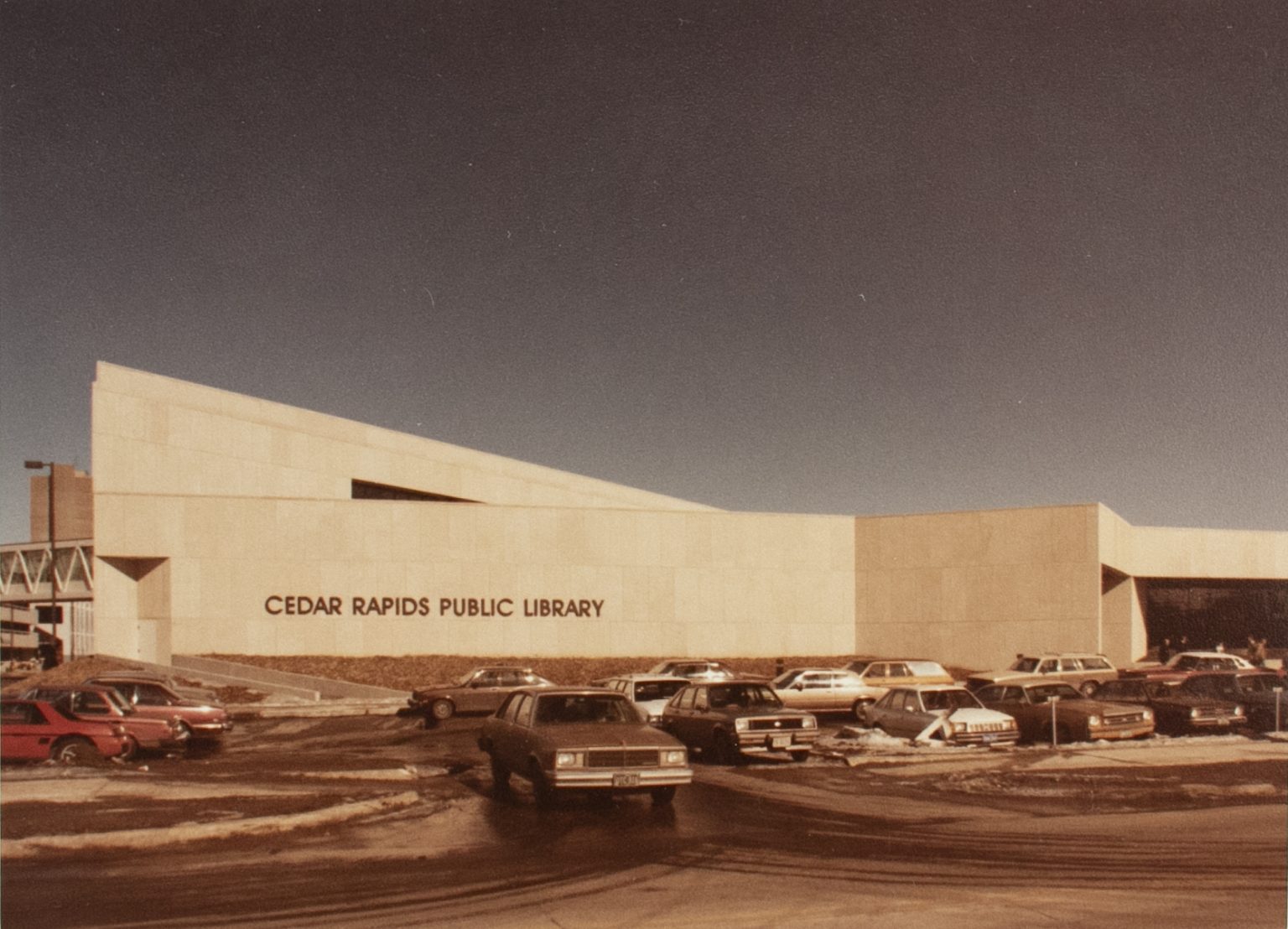 Cars sit in a parking lot in front of an angular building.