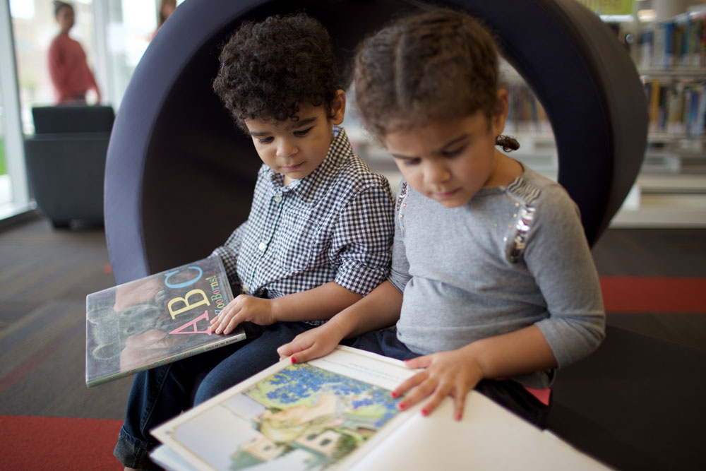 A boy and a girl sit on a bench inside the library, reading a picture book.