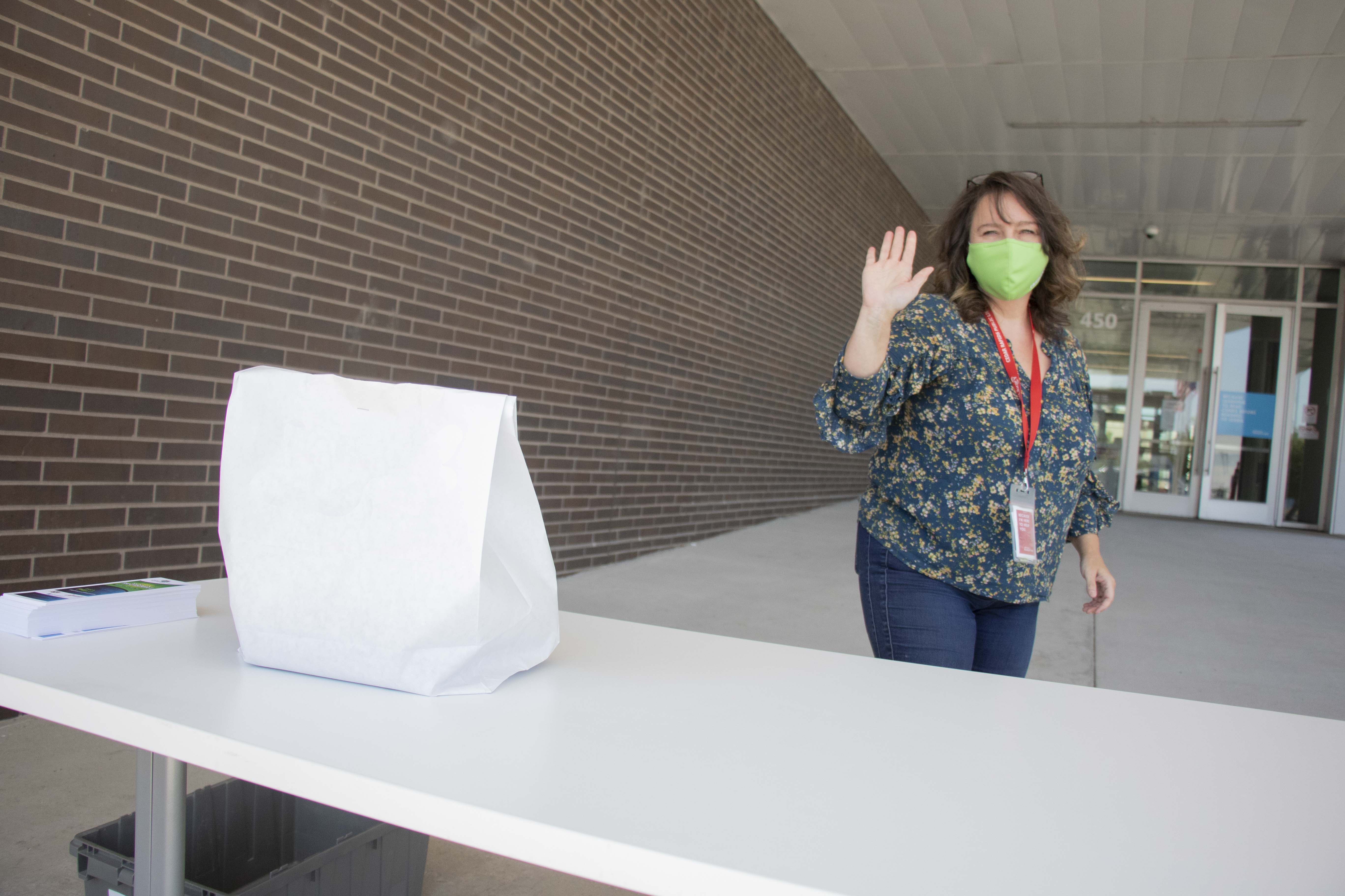 A white paper bag sits on a table while a woman in a mask waves.