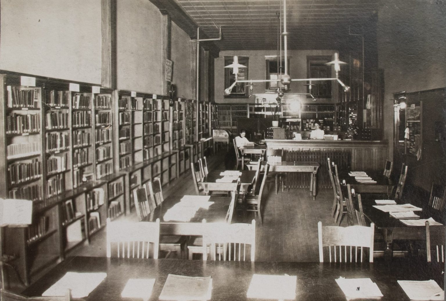 A woman sits behind a desk at the back of a room of bookshelves in this black and white photo.