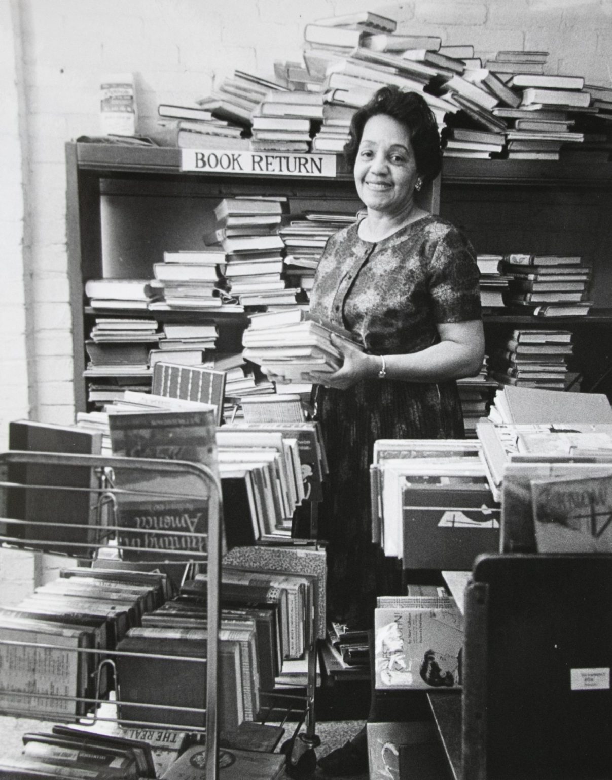 A smiling woman holds books in front of shelves of books labeled "book returns" in this black and white photo.