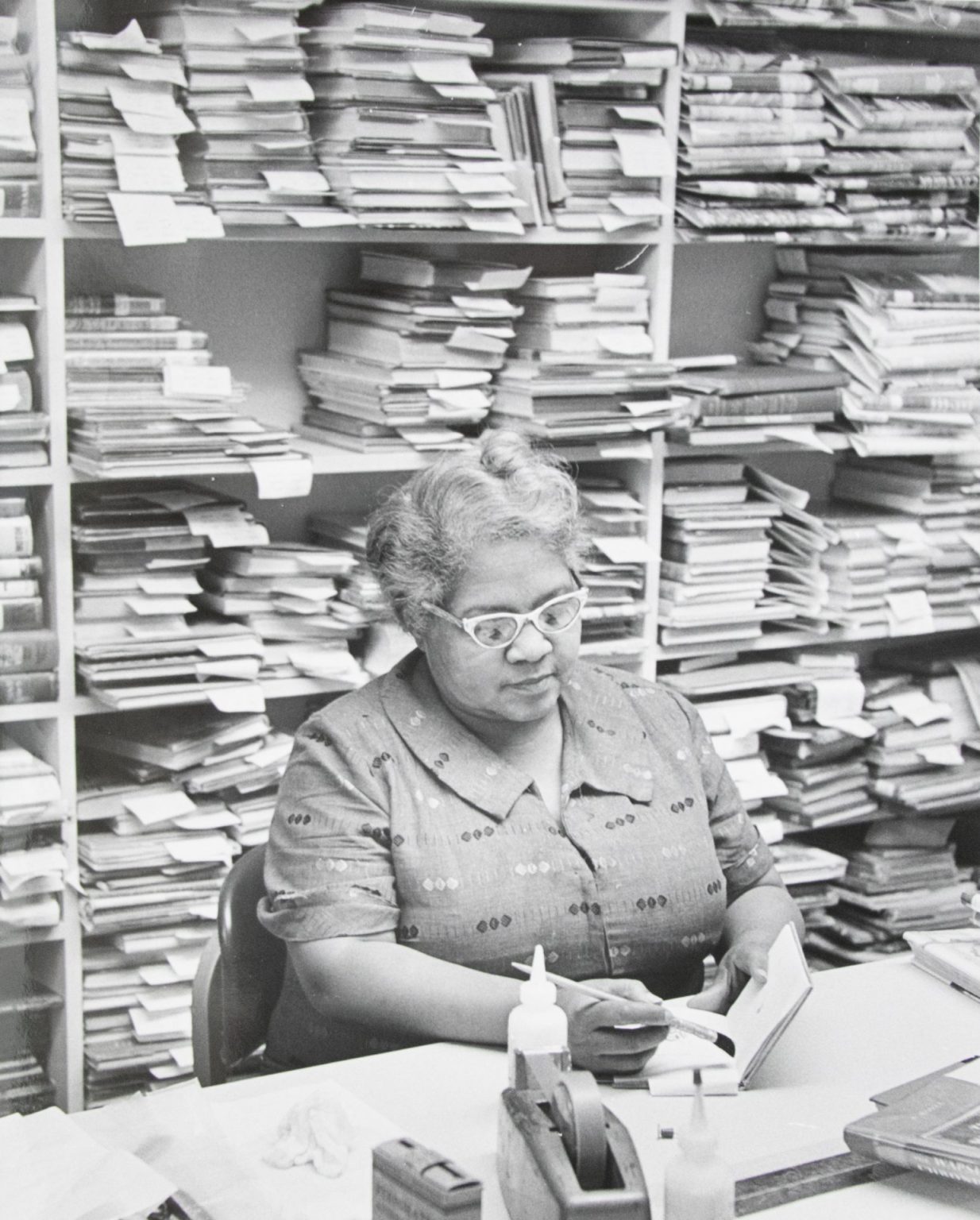 A woman with glasses uses a brush to repair a book binding in front of a shelf of books in this black and white photo.