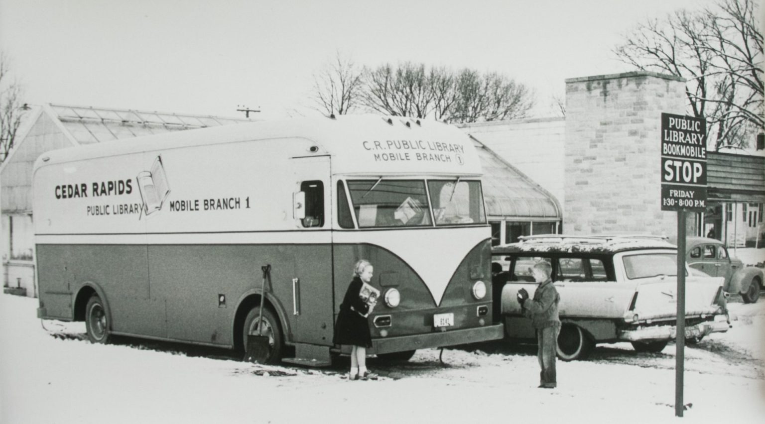 Children stand in the snow in front of a book mobile in this black and white photo.