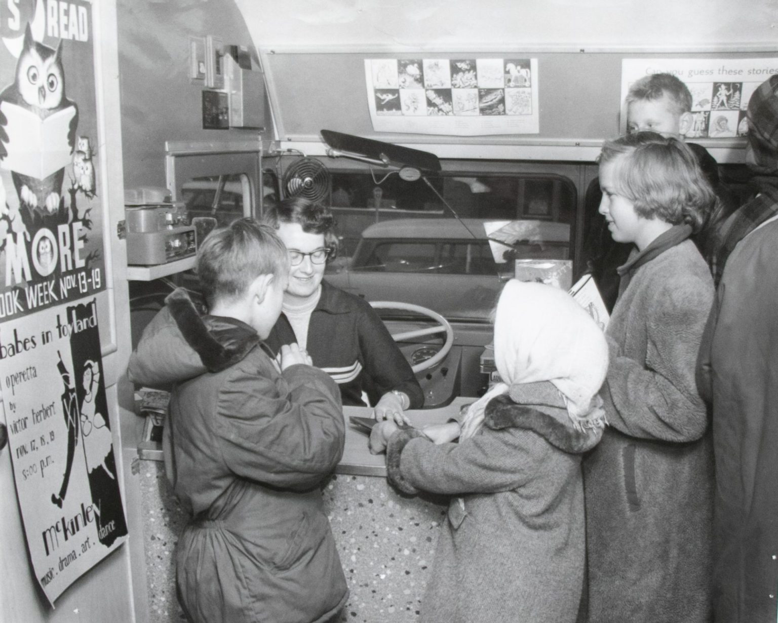 Children in winter coats gather around a check-out desk in front of a seated woman inside a book mobile in this black and white photo.