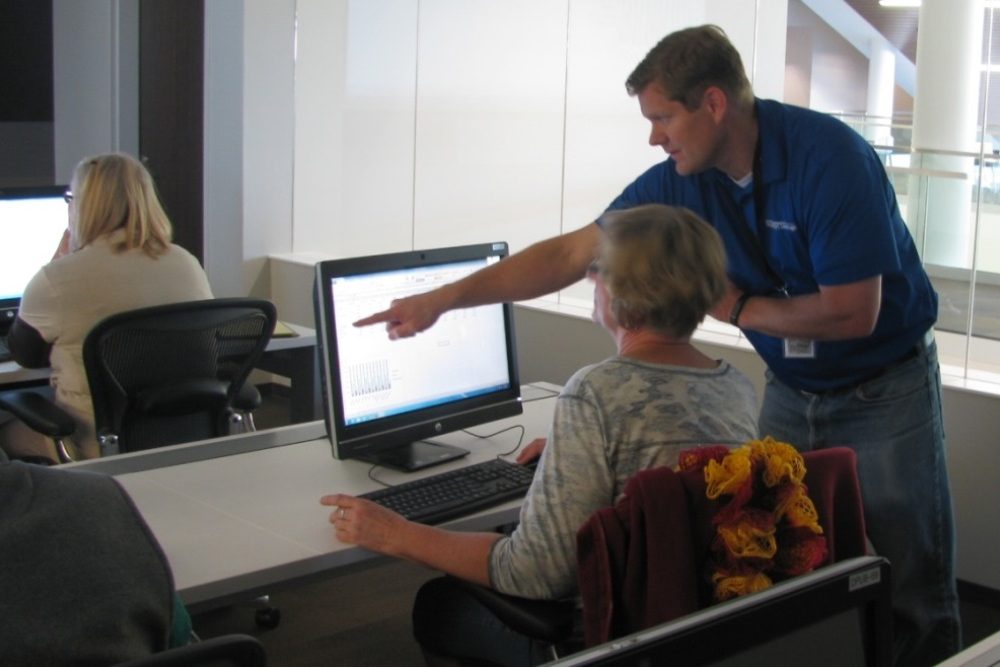A man in a blue shirt points to a computer screen while a woman sits in a chair and looks at the screen.