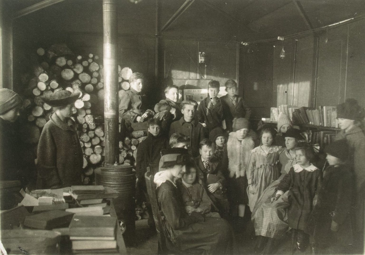 Children gather around a seated woman next to a wood stove and piles of wood in this black and white photo.