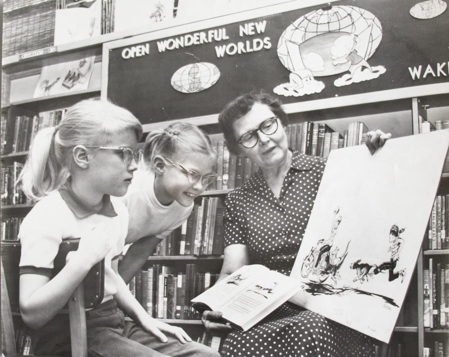 Two girls with glasses look at art held by a woman with glasses, dark hair and a polka-dotted dress in this black and white photo.