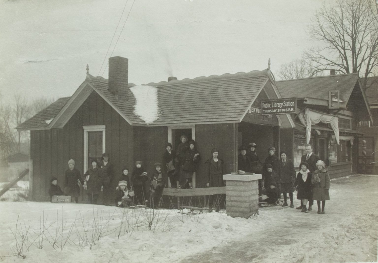 Children in winter coats stand in the snow in front of a small building, the first Kenwood branch, in this black and white photo.
