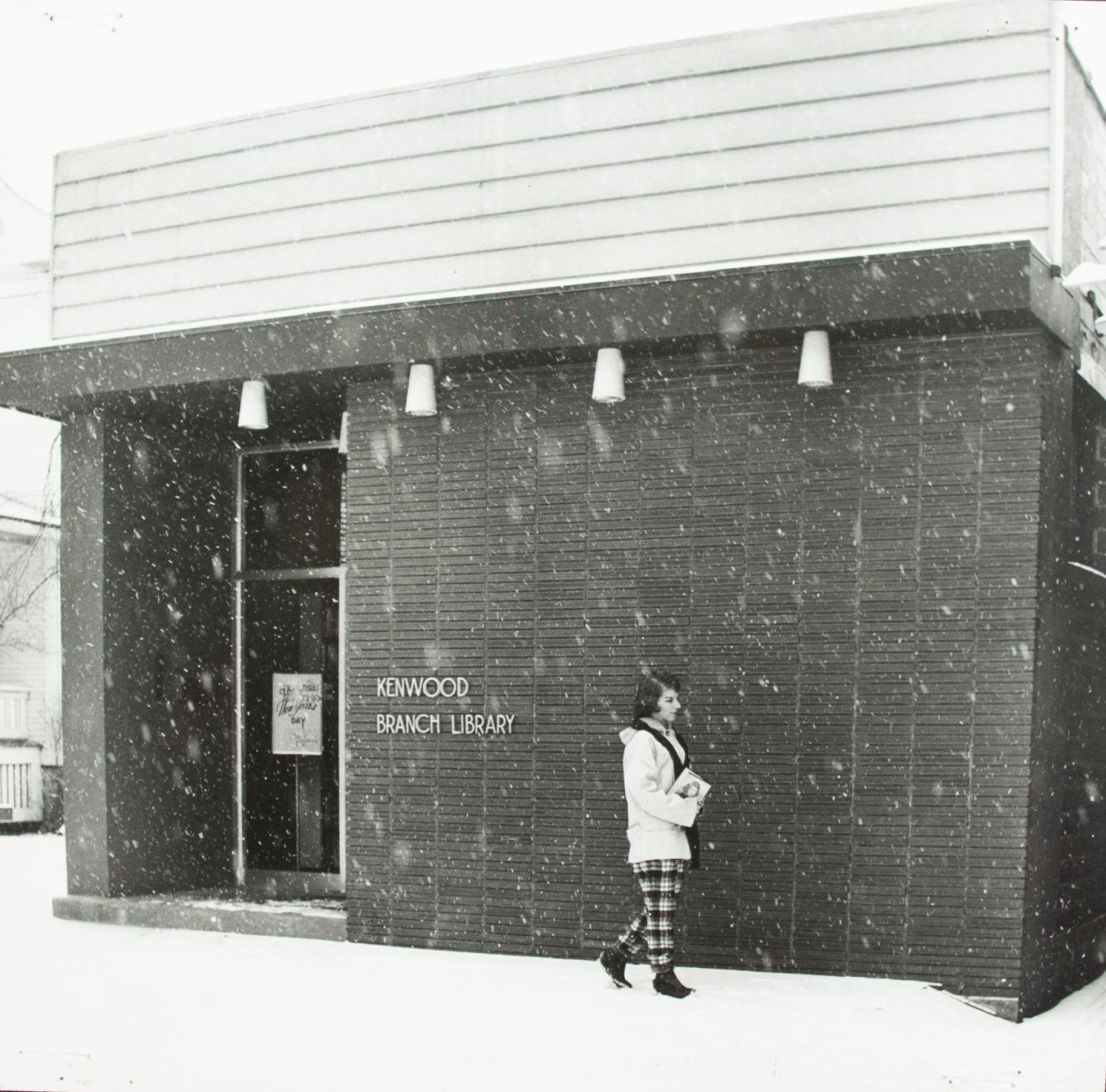 A woman in plaid pants and white coat carries a book in the snow in front of the brick Kenwood Branch.