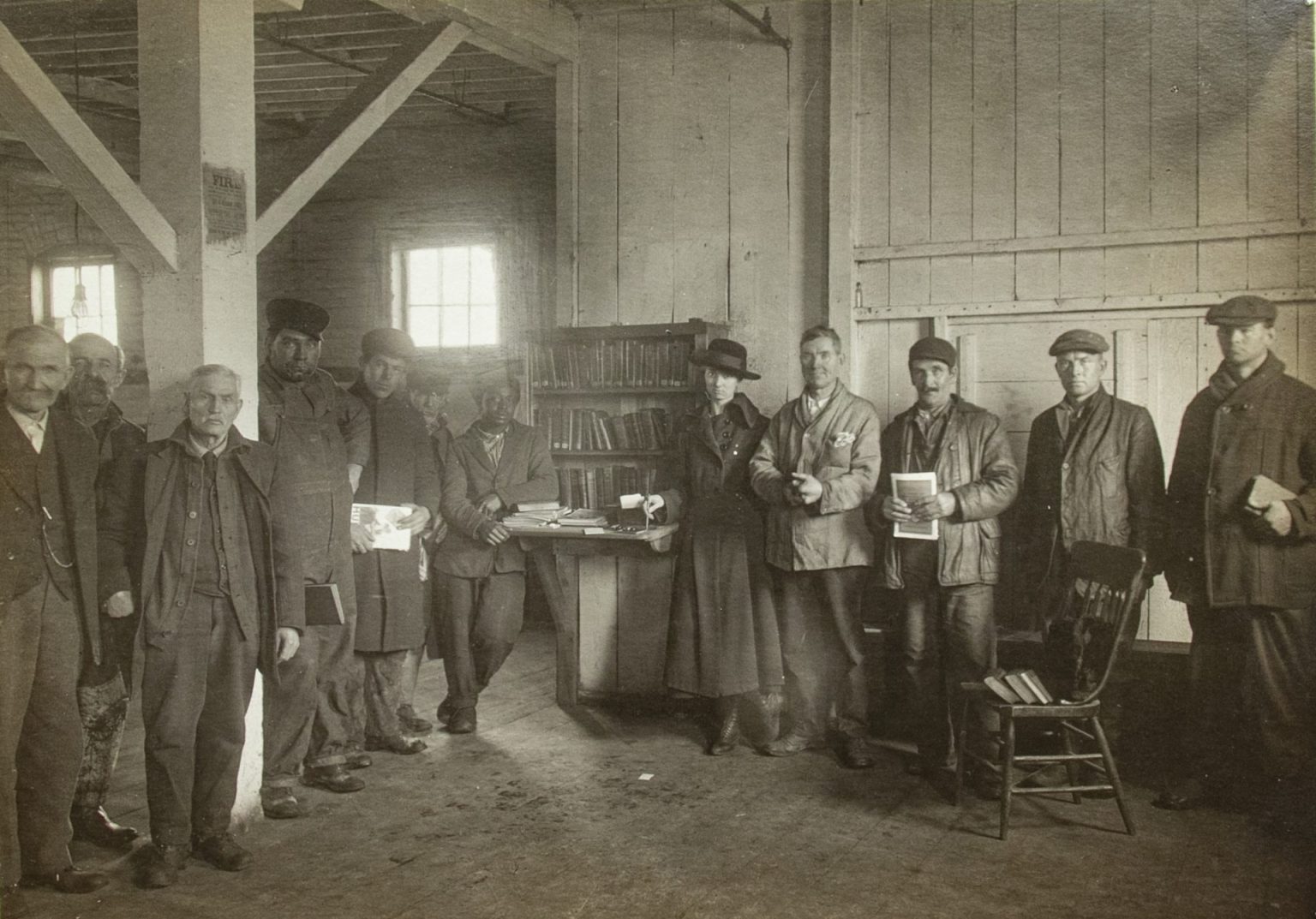 Men in work clothes stand with a woman in a dress and hat next to a book shelf in a factory room, in this black and white photo.