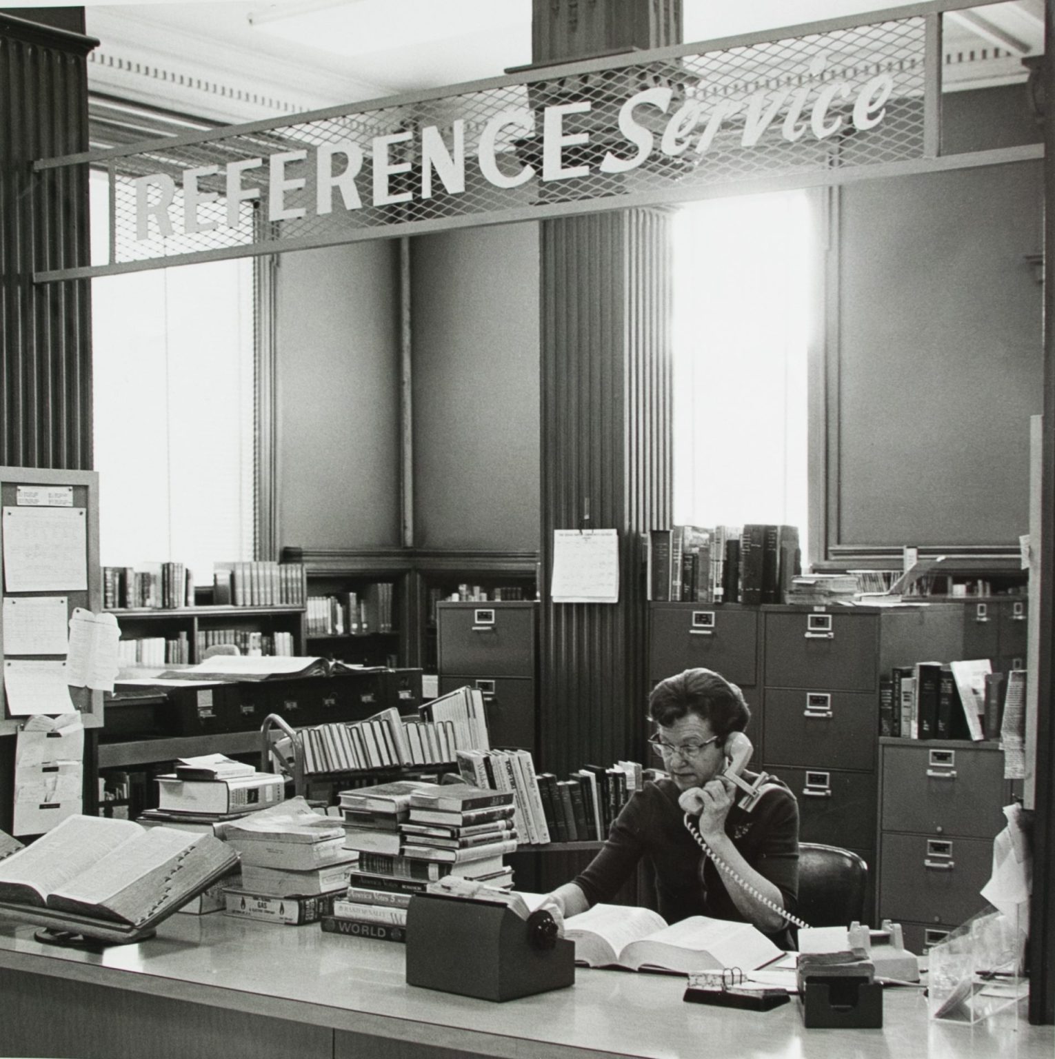 A woman answers a phone at a crowded desk under a "Reference Service" sign in this black and white photo.