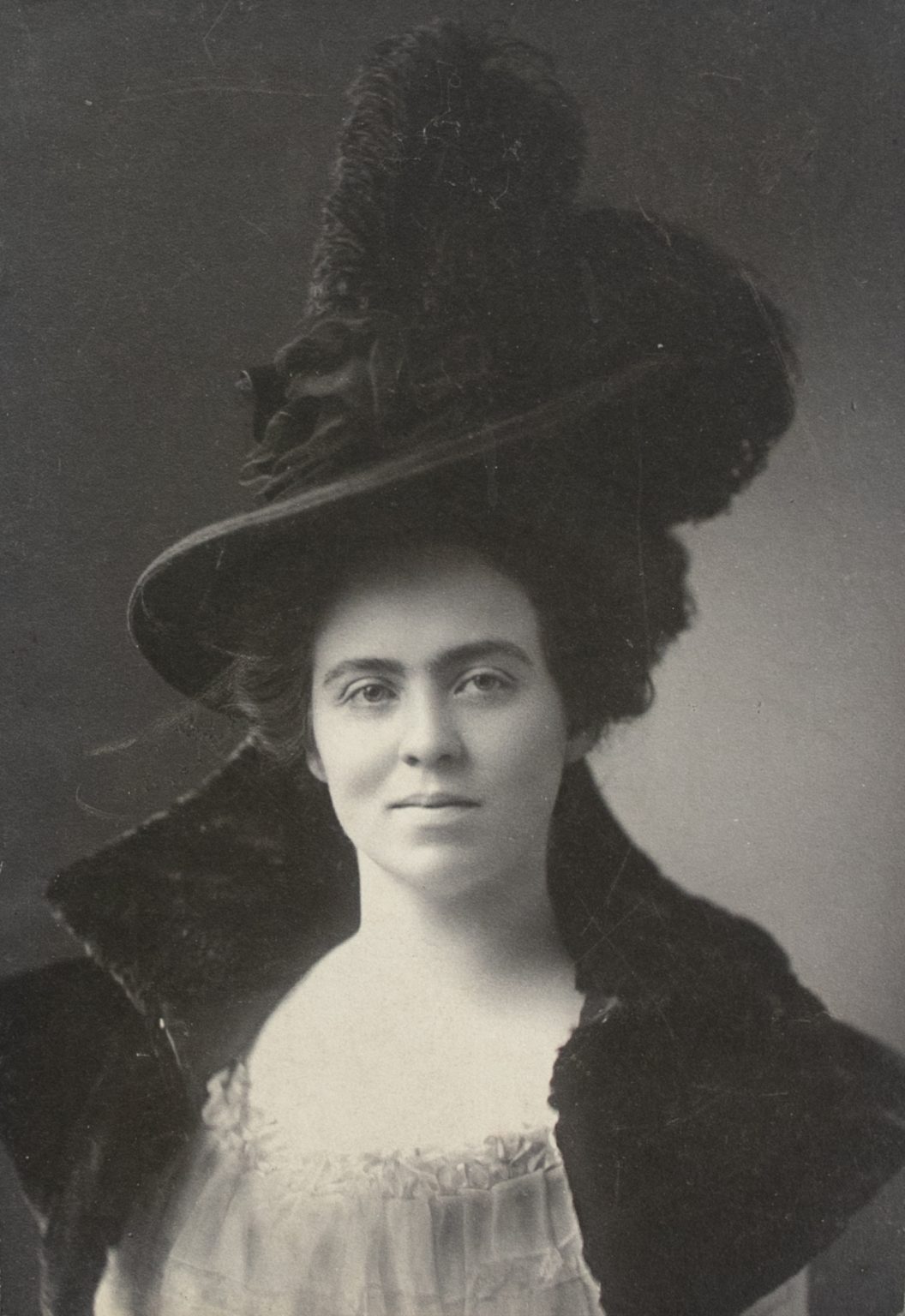 Virginia Dodge wears a feather-topped hat and fur collared-dress and looks at the camera in this black and white portrait.