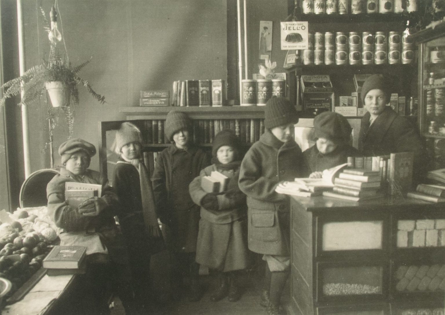 Children in winter hats and coats hold books in front of grocery store shelves in this black and white photo.
