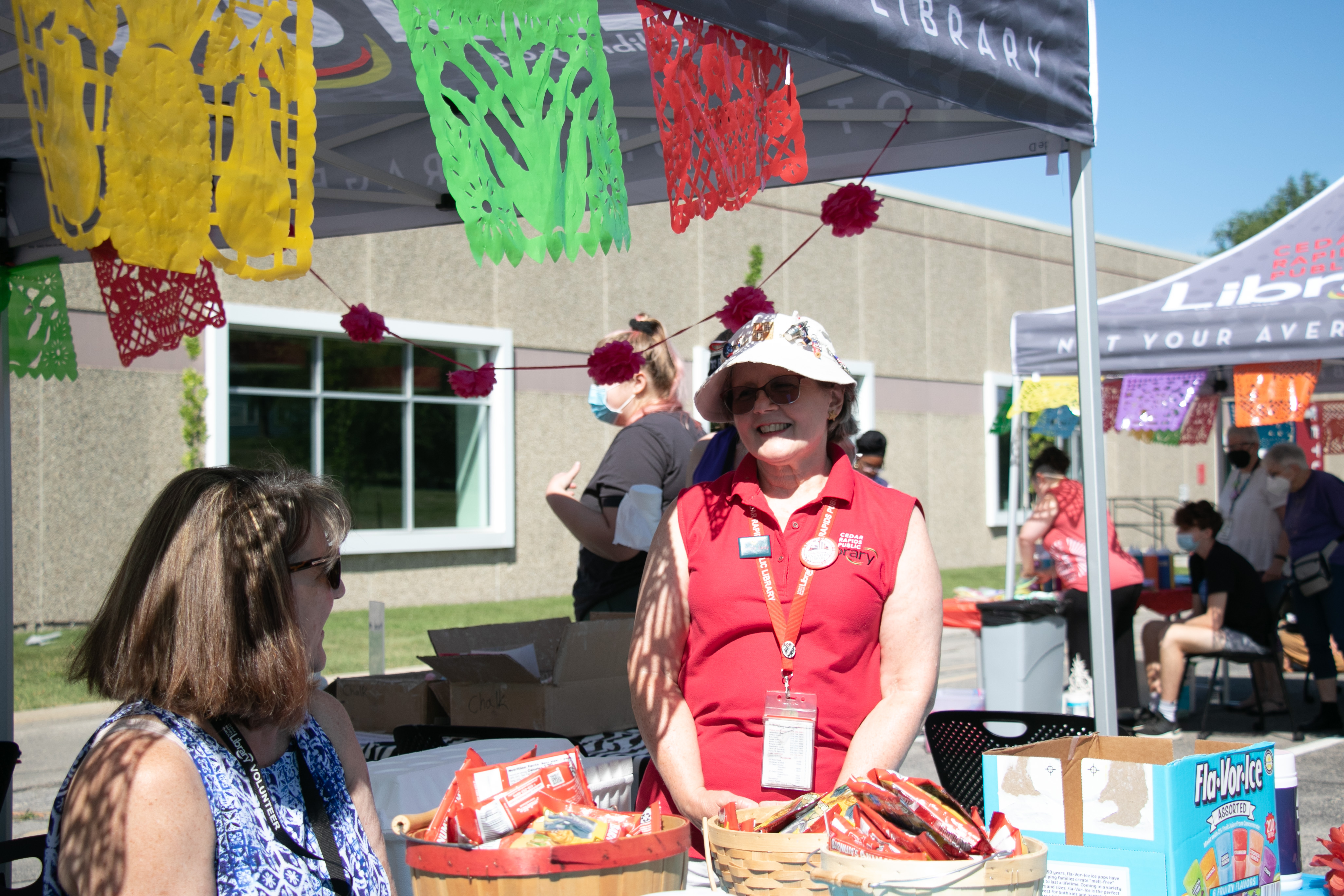 A woman in a red shirt smiles under a library tent on a sunny day.