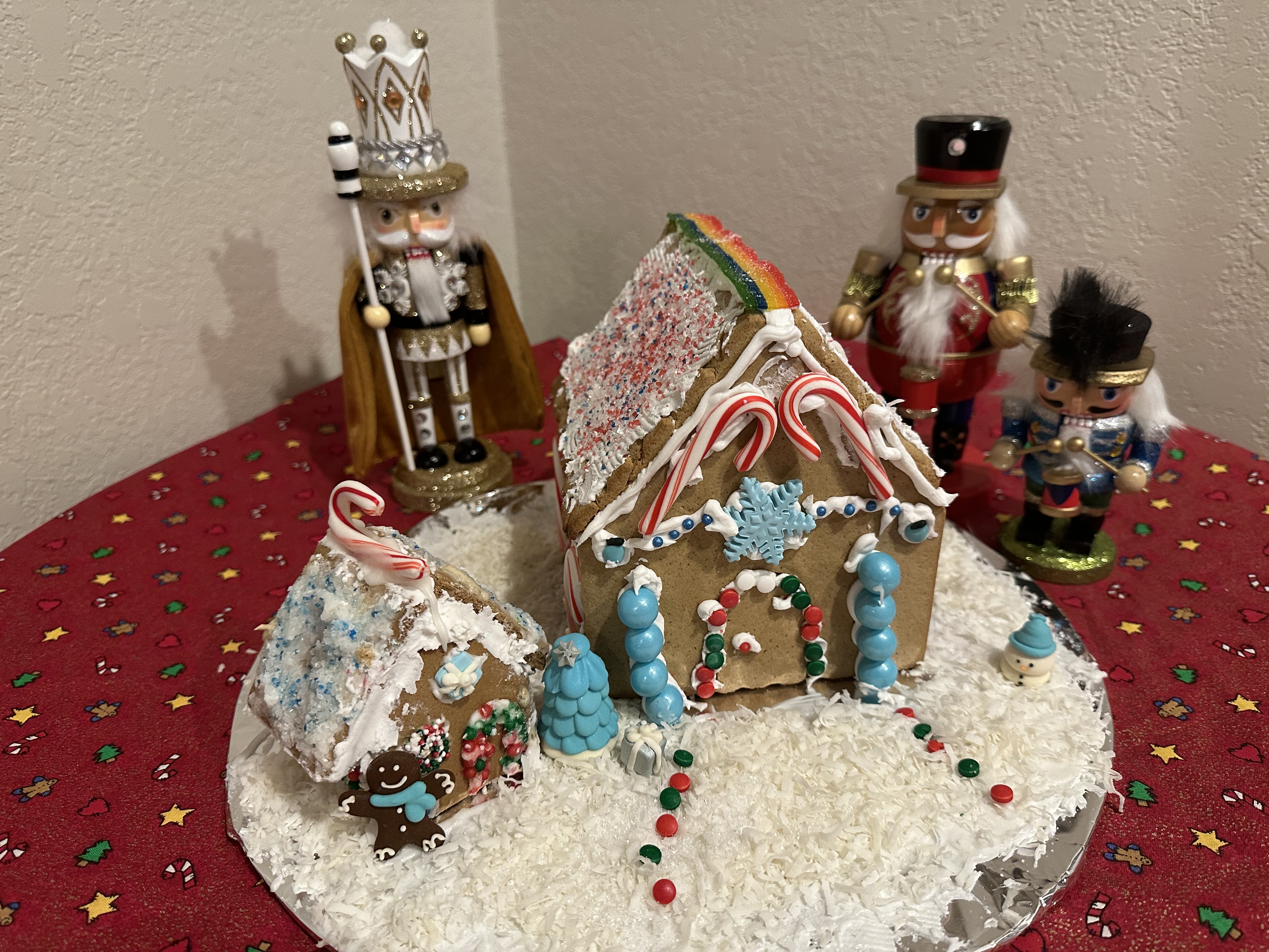 A gingerbread house and ADU sit in front of nutcrackers on a red table.