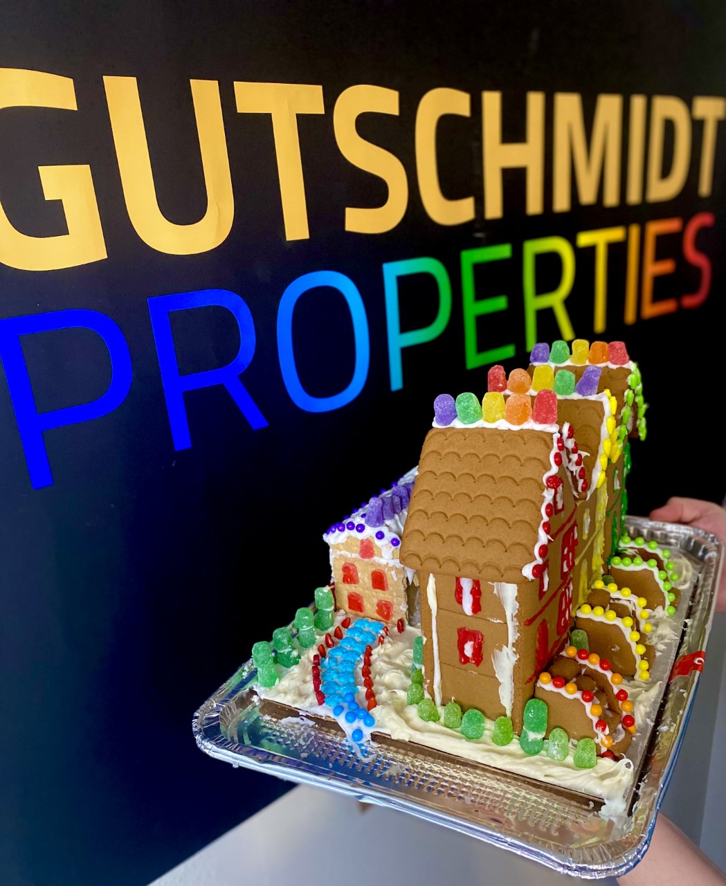 A row of gingerbread houses with rainbow icing and candy sits in front of a Gutschmidt Properties sign.