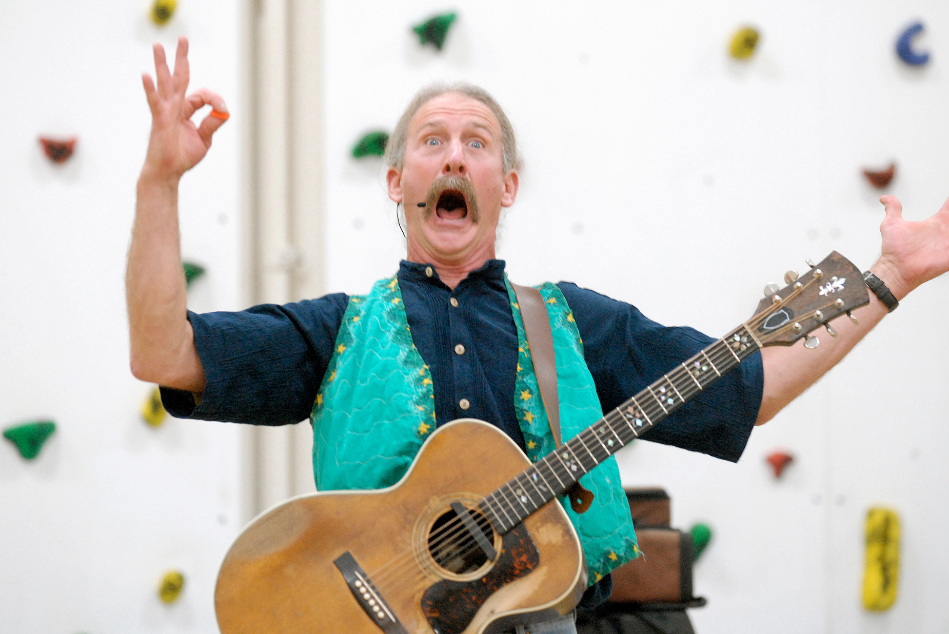 A man in a blue vest, wearing a guitar, makes an open-mouthed expression while raising his arms.