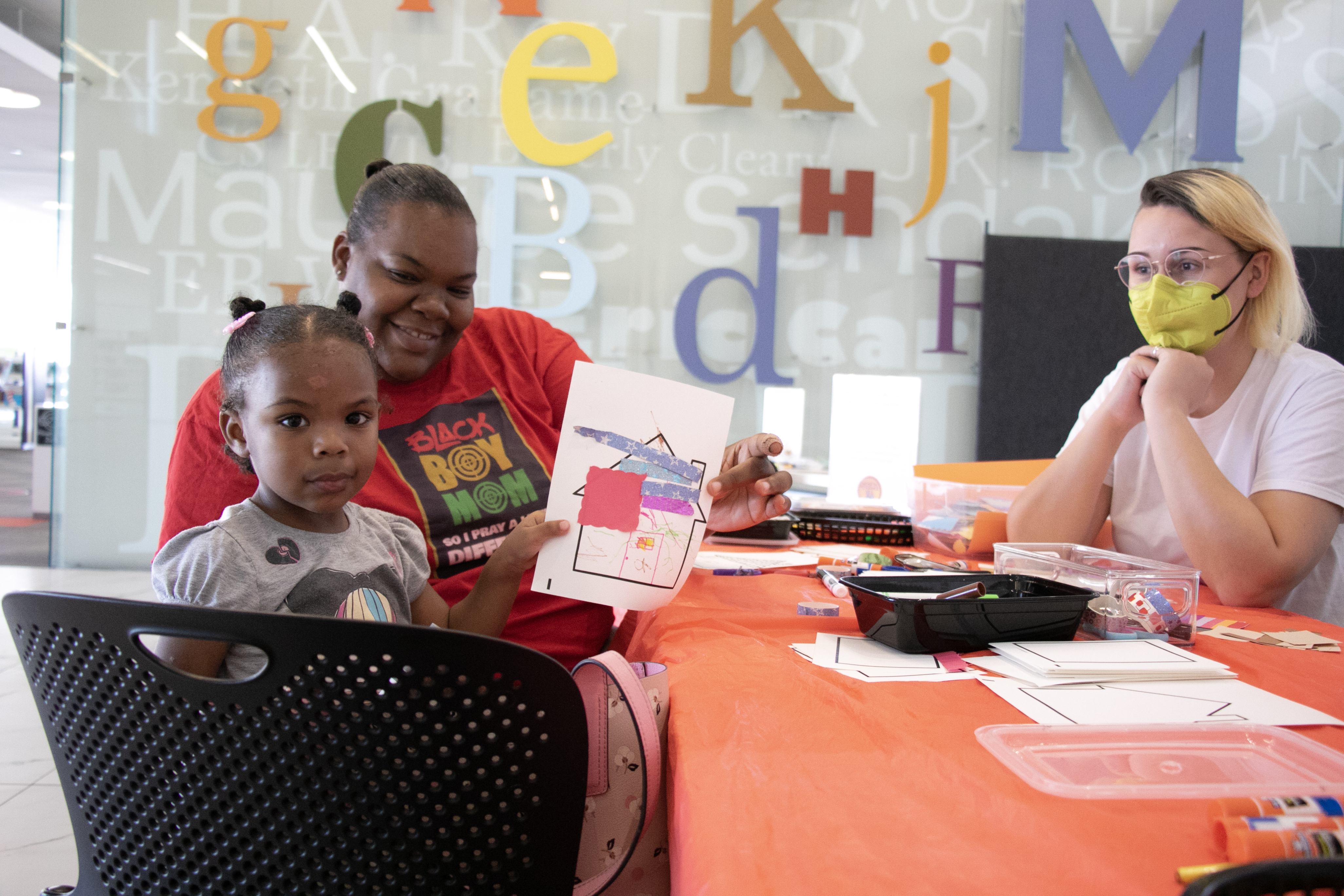 A woman and her daughter hold up their art and smile at the camera while another woman watches.