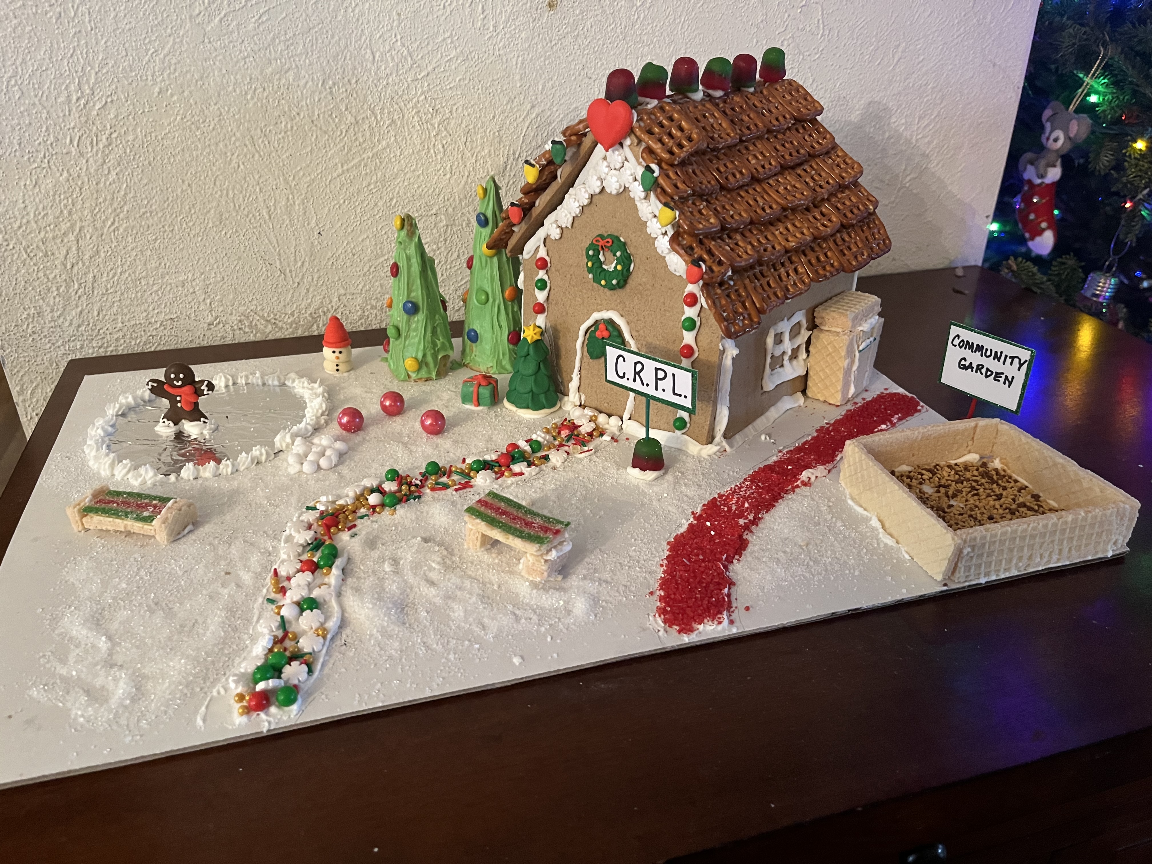 A gingerbread house has a candy path in front, a sign that says "CRPL," a pond, and a community garden.