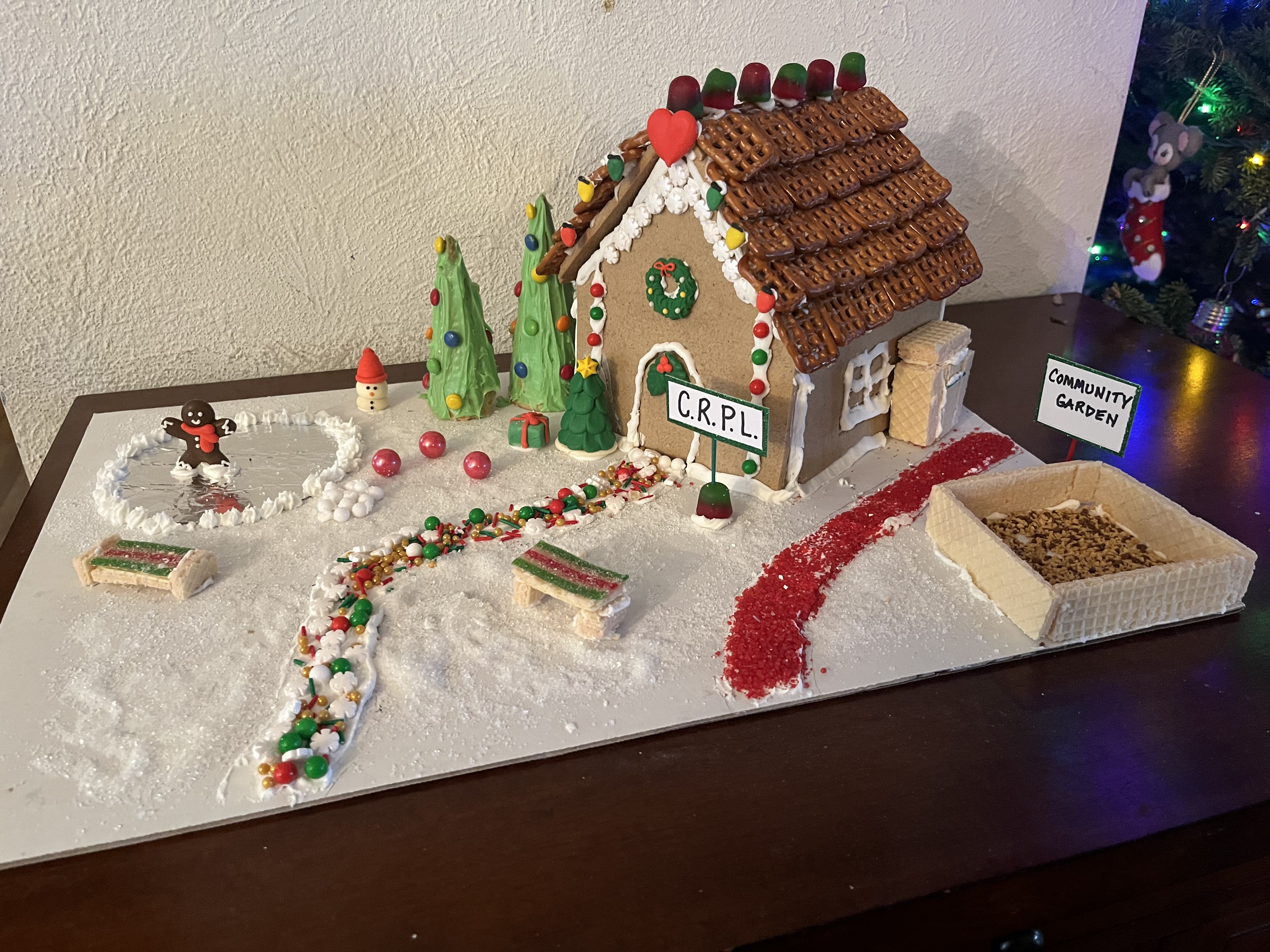 A gingerbread house has a candy path in front, a sign that says "CRPL," a pond, and a community garden.