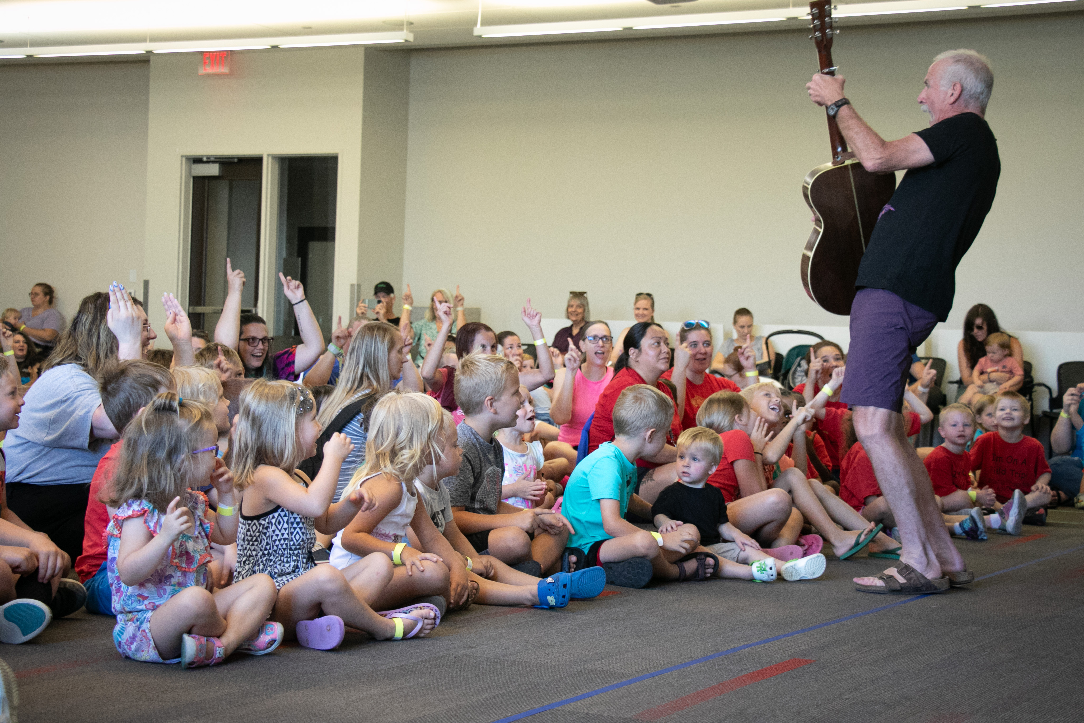 A man holds a guitar and sings in front of a crowd of seated children.