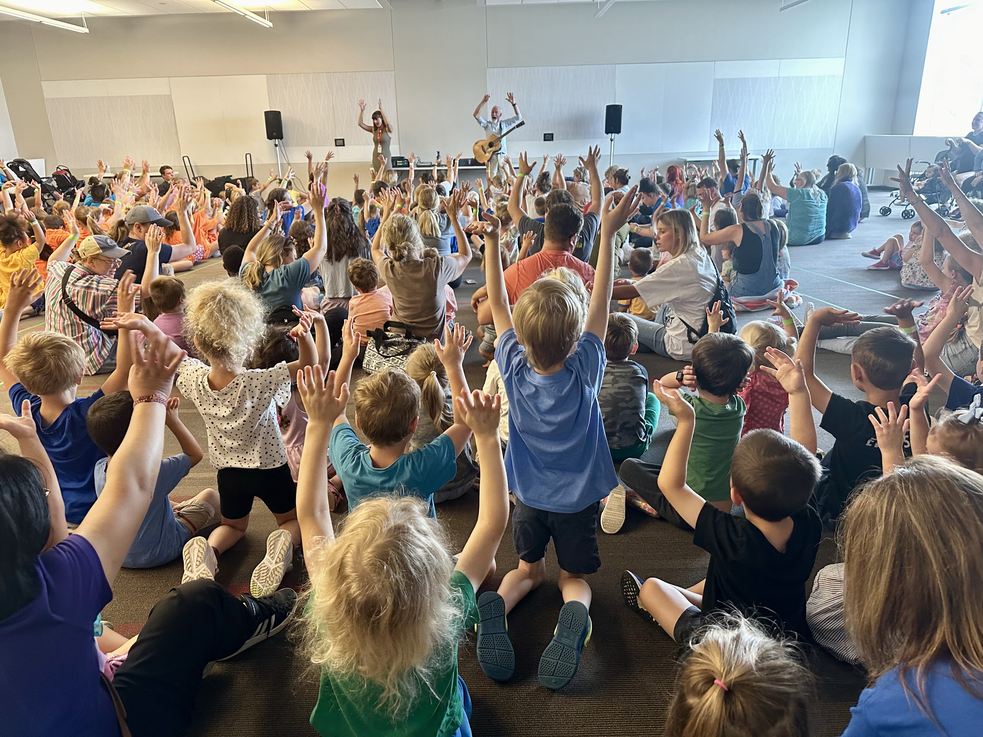 A room full of children raise their hands in the air with Tom Pease.