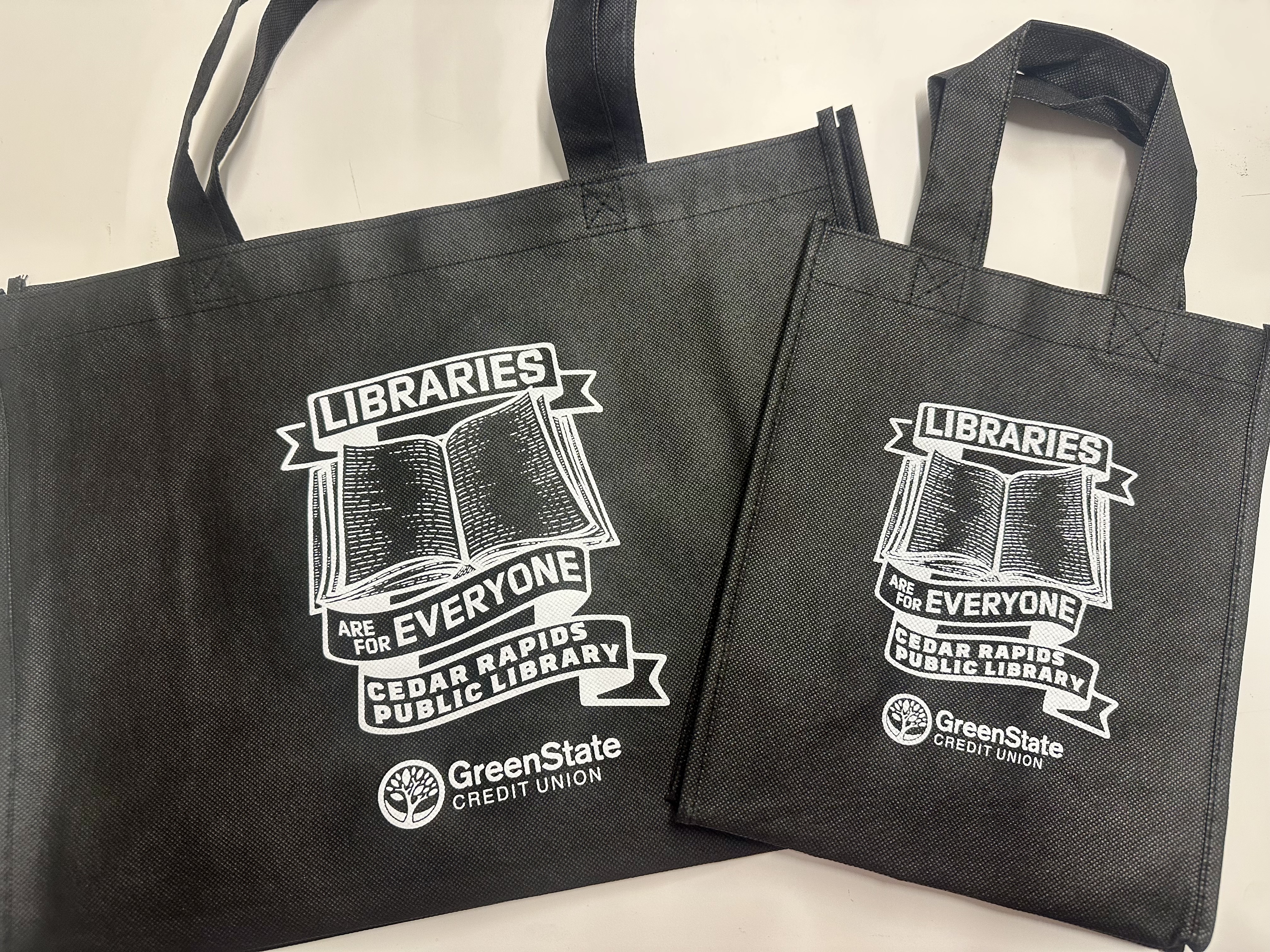 Two black tote bags say "Libraries are for Everyone" around a book illustration.