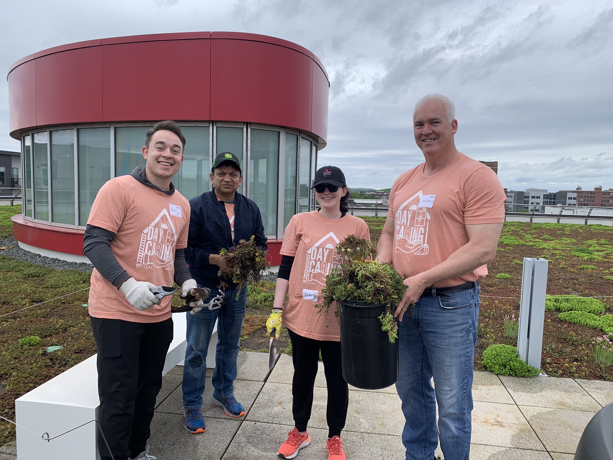 Four people in "Day of Caring" shirts hold gardening supplies on the library roof.