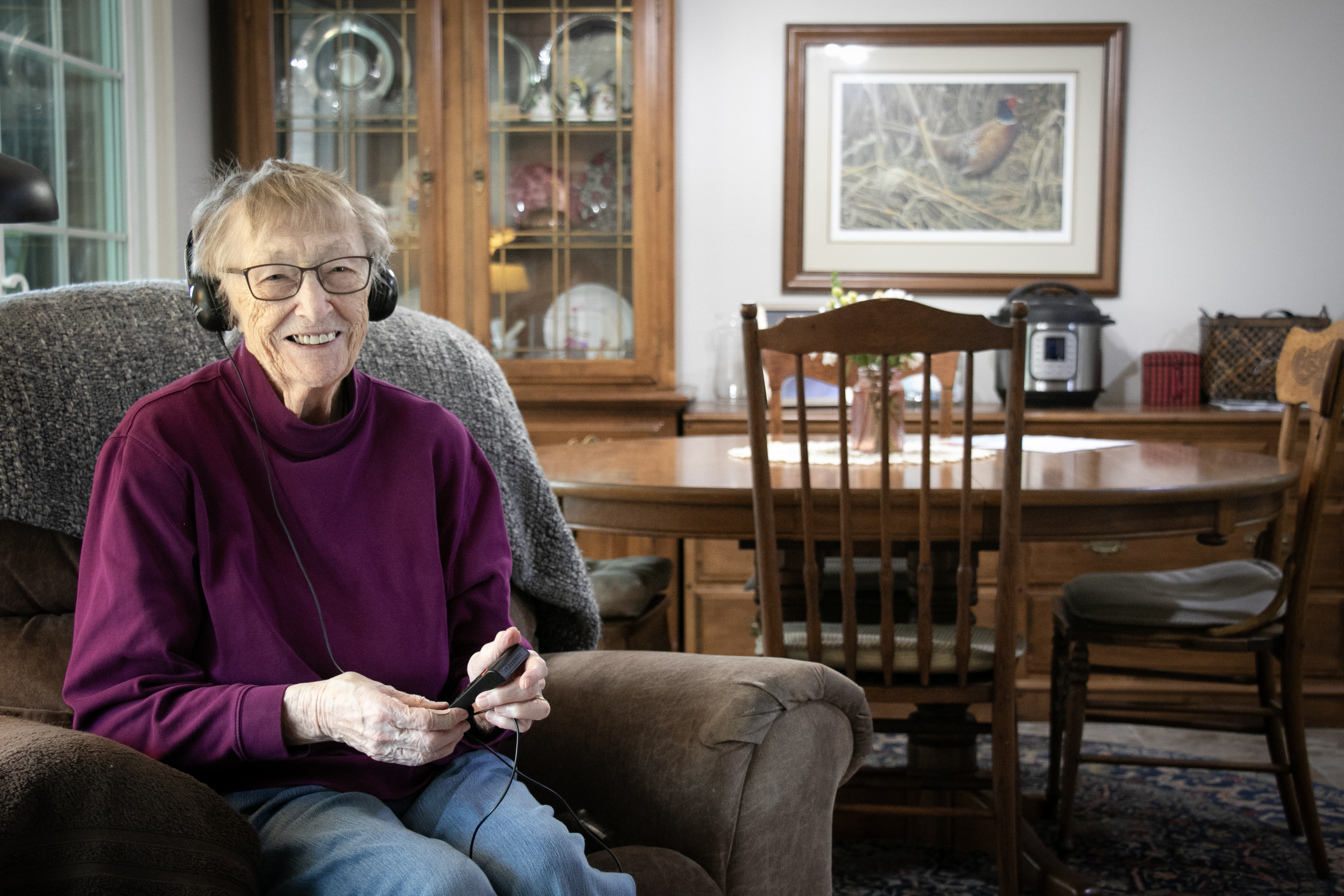 Maxine Valenta, age 93, wears headphones for an audiobook player while sitting in her living room.