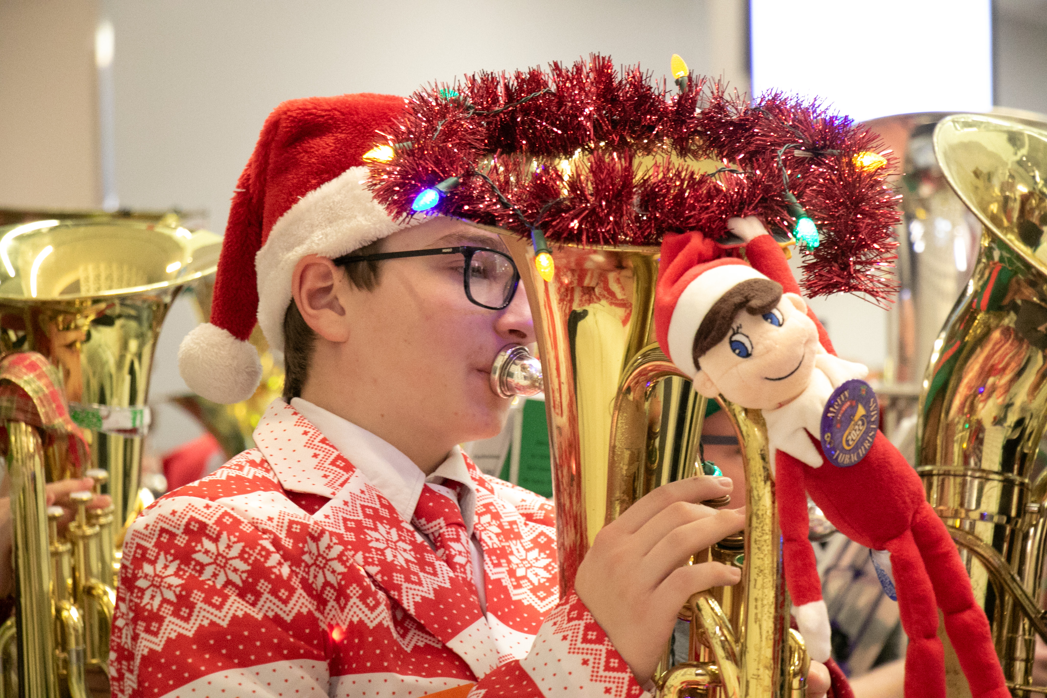 A young man wearing a Santa hat and Christmas sweater plays a tuba that is decorated with garland.