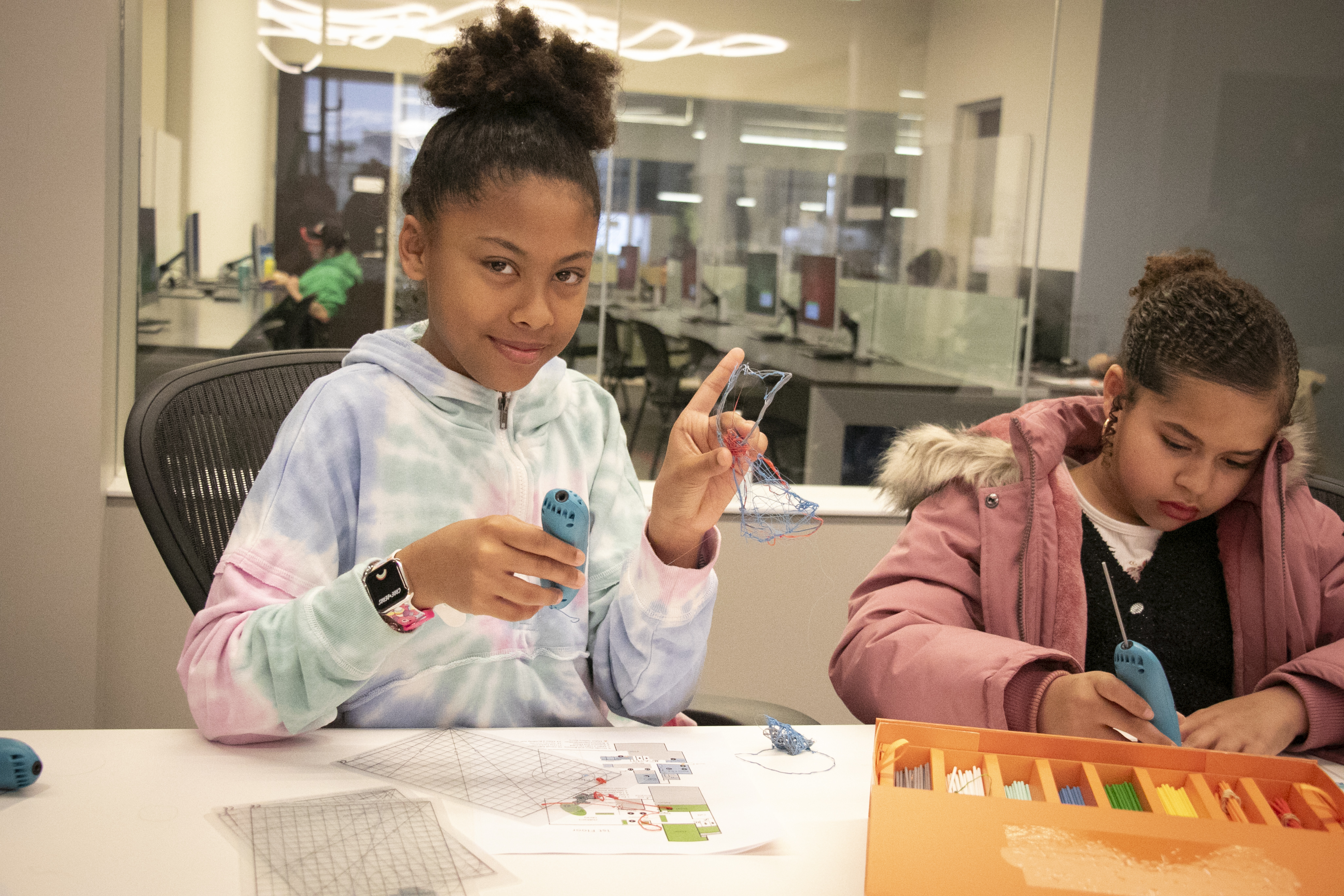 A girl smiles as she and another girl use Doodle Pens at the library.