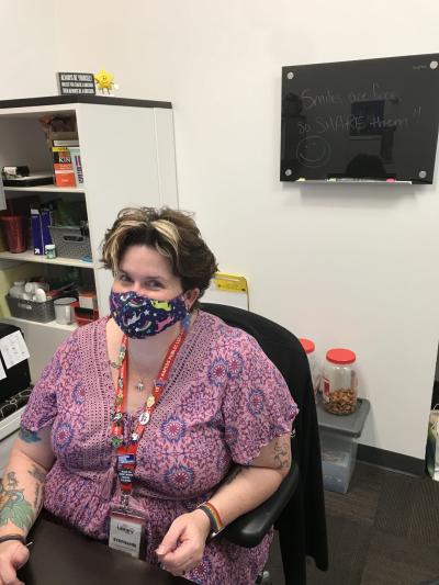 A woman in a blue face mask and purple shirt sits at a desk.