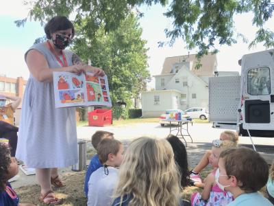 A woman in a blue dress holds up a book for a group of children to see.