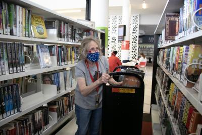 A woman wearing a face mask pushes a book cart between shelves.