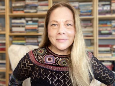 A woman with blond hair and a black patterned sweater sits in front of bookshelves.