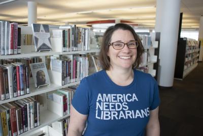 A woman in a blue t-shirt that reads "America Needs Librarians" smiles in front of book shelves.