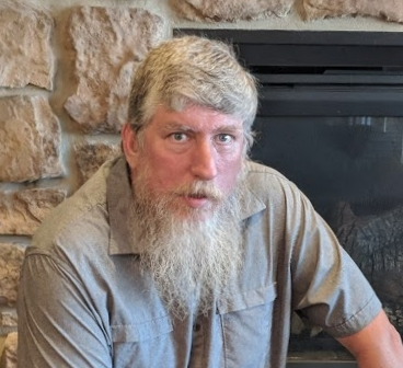 Joseph Engler has a long beard and white hair and sits in front of a fireplace.