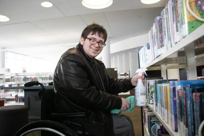 A man in a wheelchair smiles at the camera while holding cleaning supplies in front of bookshelves.
