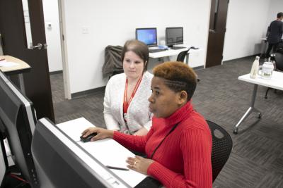 A woman in a red sweater looks at a computer while another woman helps her.