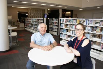 An older man and a woman smile at the camera in front of book shelves in the library.