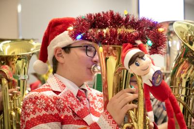 A young man wearing a Santa hat and Christmas sweater plays a tuba that is decorated with garland.