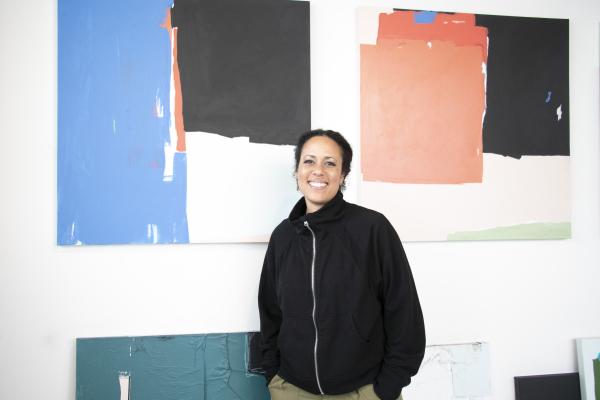 A woman, Akwi Nji, stands in front of her abstract paintings featuring blocks of color.