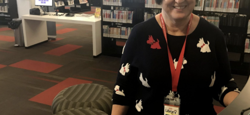 A woman with a black sweater with a dog pattern smiles in front of book shelves