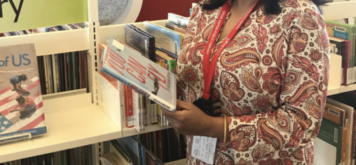 A woman in a red shirt holds a book in front of shelves.