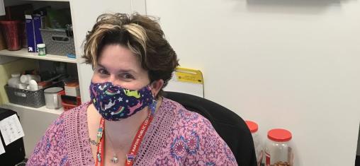 A woman in a blue face mask and purple shirt sits at a desk.