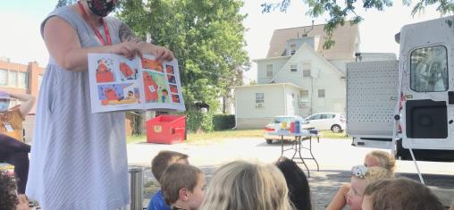 A woman in a blue dress holds up a book for a group of children to see.