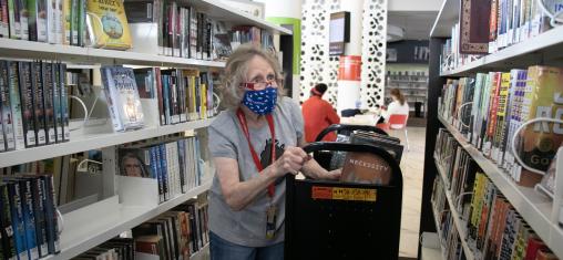 A woman wearing a face mask pushes a book cart between shelves.